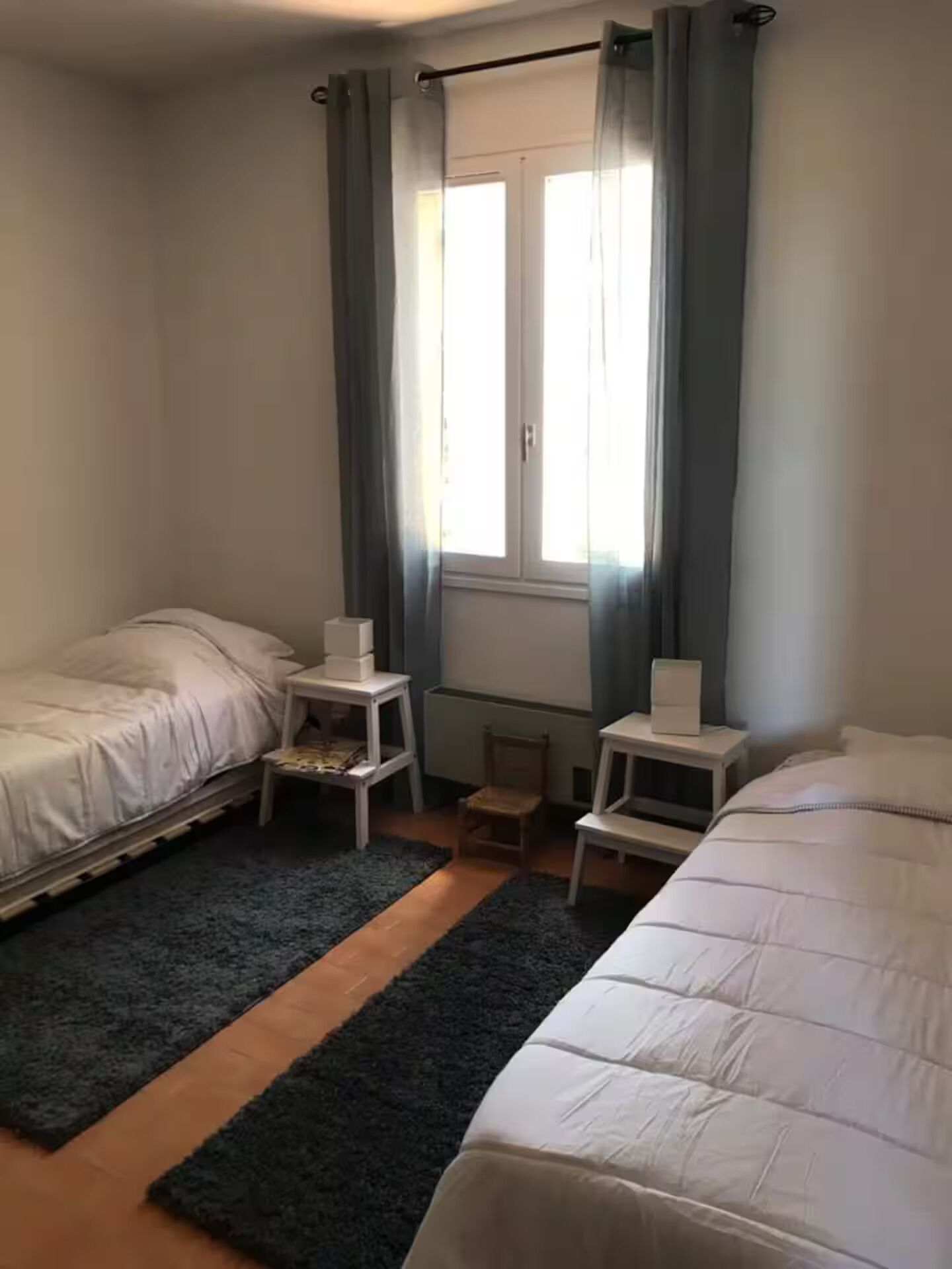 Un dormitorio con dos camas, una ventana con cortinas, un radiador y una mesita de noche con lámpara.