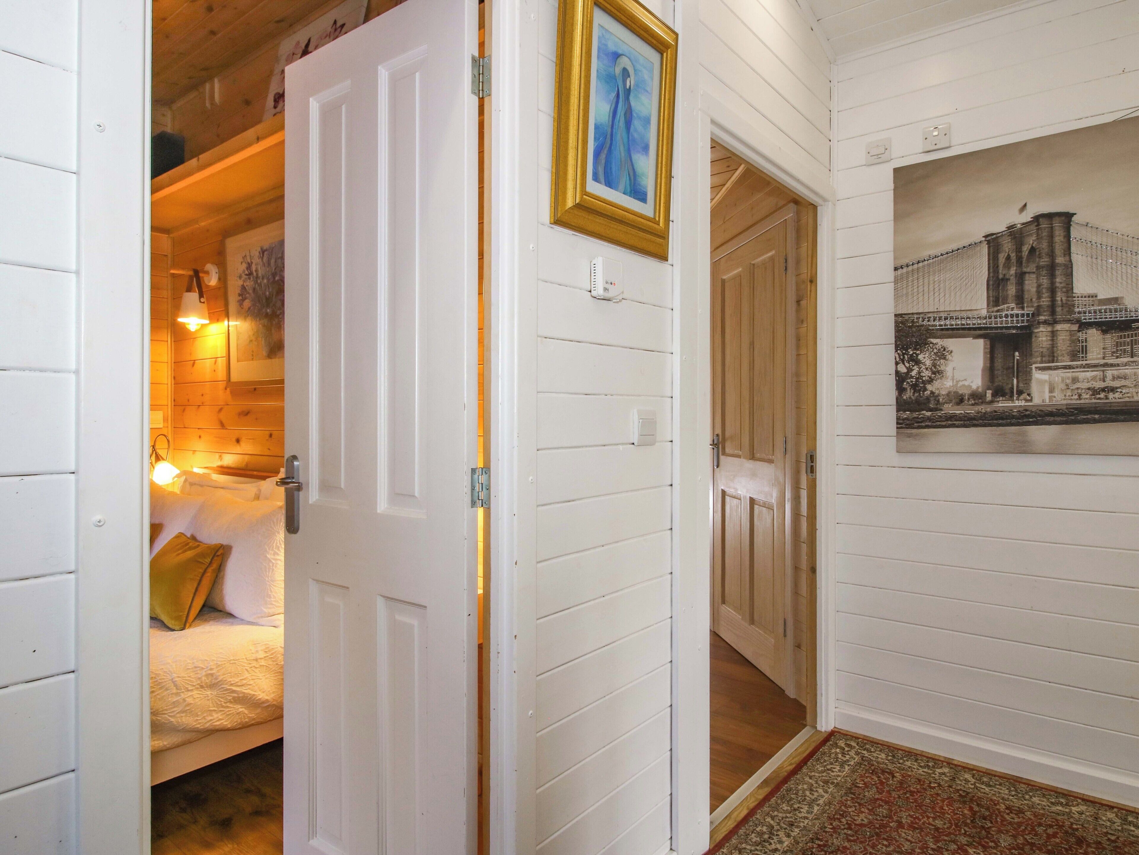 A hallway with white paneled walls, a bed with a yellow pillow, a framed picture, and a door leading to another room.