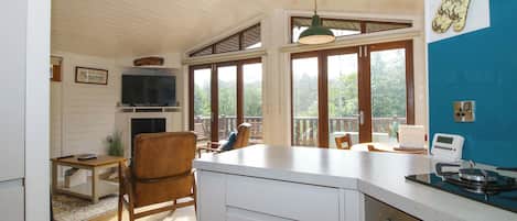 A kitchen with a TV, wooden chairs, a dining table, and a view through large windows.