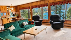 Een gezellig cabinteinterieur met een groene bank, twee fauteuils, een houten salontafel en grote ramen met uitzicht op bomen en een meer.