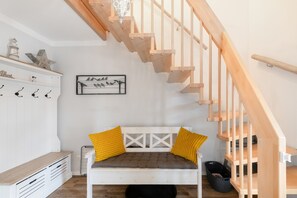 Une pièce avec un escalier en bois, un banc blanc agrémenté de coussins jaunes, une sculpture d’oiseau fixée au mur et un meuble blanc muni de crochets.