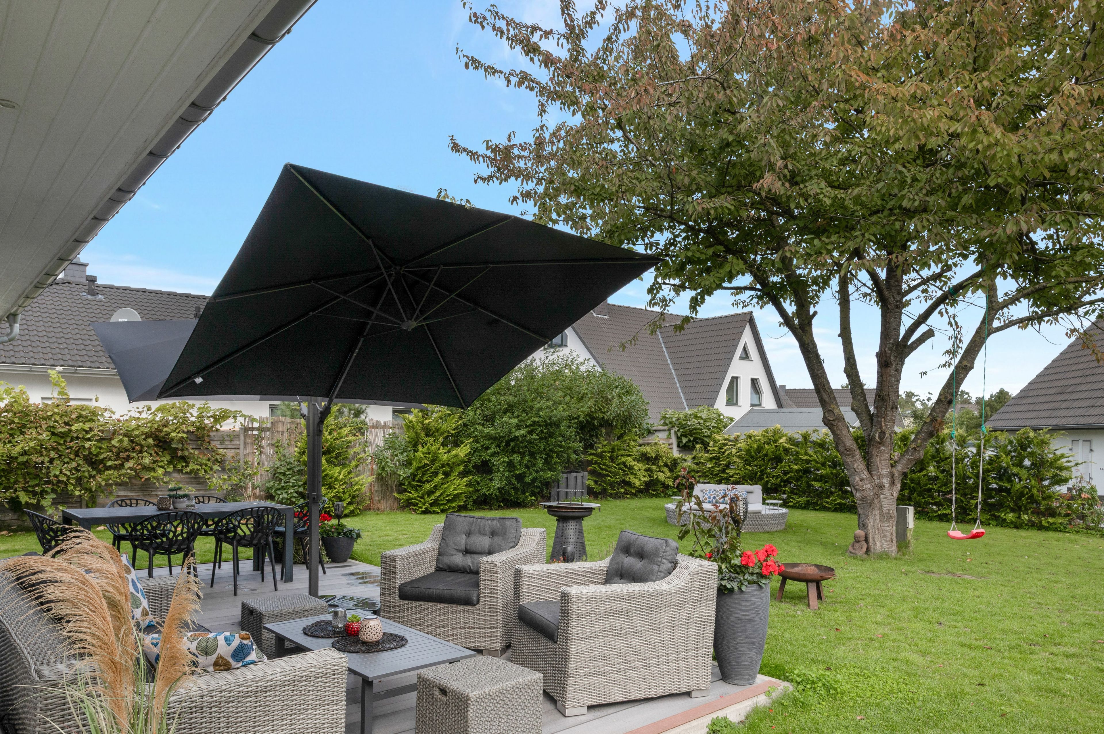 An outdoor patio area with wicker furniture, a large umbrella, and a garden with a tree and a swing.