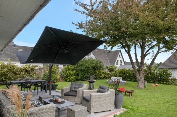 Ein möblierter Terrassenbereich mit Korbmöbeln, einem großen Sonnenschirm und einem Garten mit Baum und Schaukel.