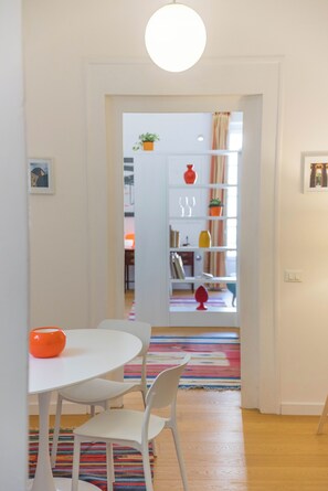 A room with a white table, two chairs, a colorful rug, a white bookshelf, and a door leading to another room. - Adenzia Apartment (Palermo)