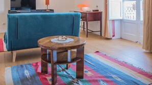 A living room with a blue sofa, a small wooden table, a TV, and a window with curtains. - Adenzia Apartment (Palermo)