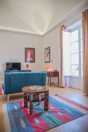 A living room with a blue sofa, a small wooden table, a TV, and a window with curtains. - Adenzia Apartment (Palermo)
