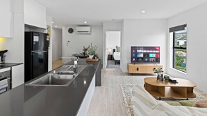 A modern kitchen with a central island, a dining area with a TV, and a bedroom visible through an open door.
