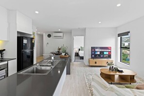A modern kitchen with a central island, a dining area with a TV, and a bedroom visible through an open door. - Stylish 2BR Near Garden City & Sunnybank Food Hub (Upper Mount Gravatt)