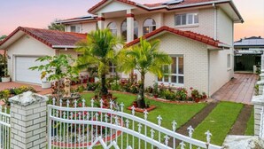 Une maison à deux étages, avec un toit recouvert de tuiles rouges, une clôture blanche et un jardin bien entretenu agrémenté de palmiers et de fleurs.