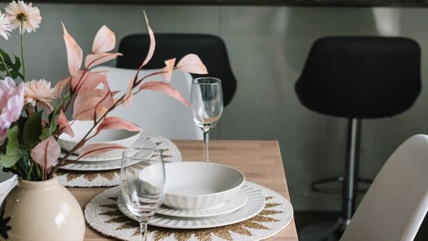 Ein Esstisch ist gedeckt mit weißen Tellern, Gläsern und einer Vase mit Blumen.