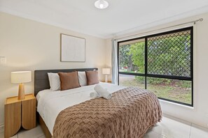 Ein Schlafzimmer mit Bett, Nachttisch, Fenster mit Blick auf Grünflächen und Deckenleuchte.
