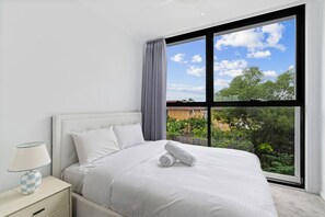Ein Schlafzimmer mit großem Fenster, einem Bett mit weißer Bettwäsche, einem Nachttisch mit Lampe und Blick auf Grünflächen und Gebäude.