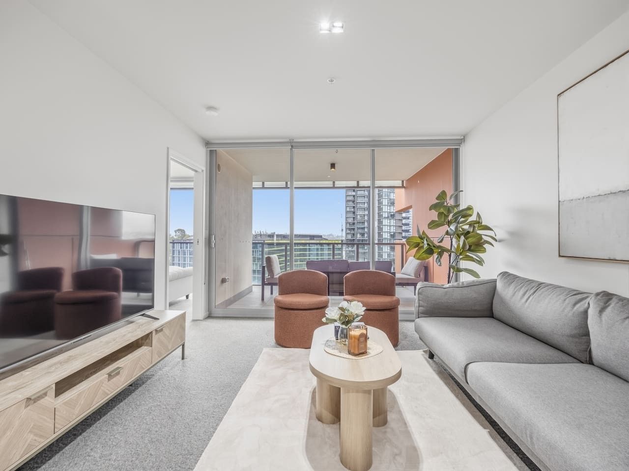A modern living room with a flat-screen TV, a grey sofa, two armchairs, a coffee table, and a large window offering a city view.