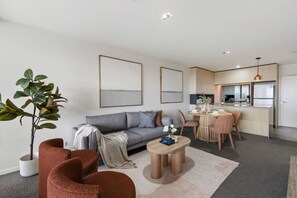 Een moderne woonkamer met een grijze bank, een salontafel en een eethoek met een tafel en stoelen. Er is een keuken met kasten en een koelkast.