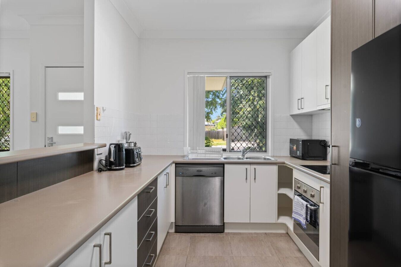 A modern kitchen with white cabinets, a dishwasher, a refrigerator, and a window with a view of greenery.