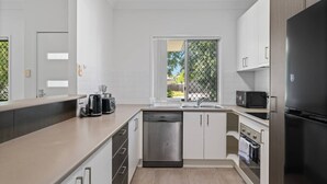 Una cocina moderna con armarios blancos, lavavajillas, refrigerador y ventana con vista a la vegetación.