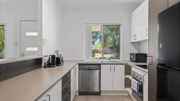 A modern kitchen with white cabinets, a dishwasher, a refrigerator, and a window with a view of greenery.