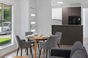 A dining area with a round table set for four, surrounded by grey upholstered chairs. A kitchen with dark cabinets and a black refrigerator is visible in the background. - Comfort Nest|2BR Quiet Comfort Retreat for Relax (Mount Warren Park)