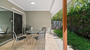 Un patio con una mesa, sillas, un vidrio y vistas de un jardín con cerco.