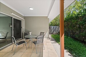 A patio with a table and chairs, a glass door, and a view of a garden with a fence. - Comfort Nest|2BR Quiet Comfort Retreat for Relax (Mount Warren Park)