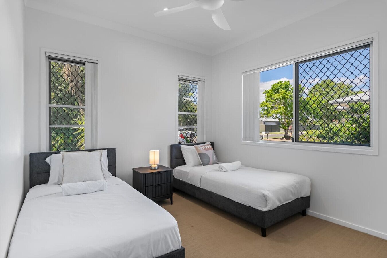 A bedroom with two beds, a nightstand, a ceiling fan, and large windows with a view of greenery.