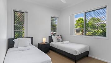 A bedroom with two beds, a nightstand, a ceiling fan, and large windows with a view of greenery.