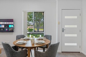 Un comedor con una mesa redonda dispuesta para cuatro personas, un televisor montado en la pared mostrando diferentes canales, y una puerta con vista al exterior.