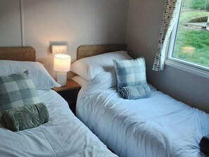 A bedroom with two beds, a bedside lamp, a window with curtains, and a power outlet on the wall. - Josephine Nb37 (Newquay)