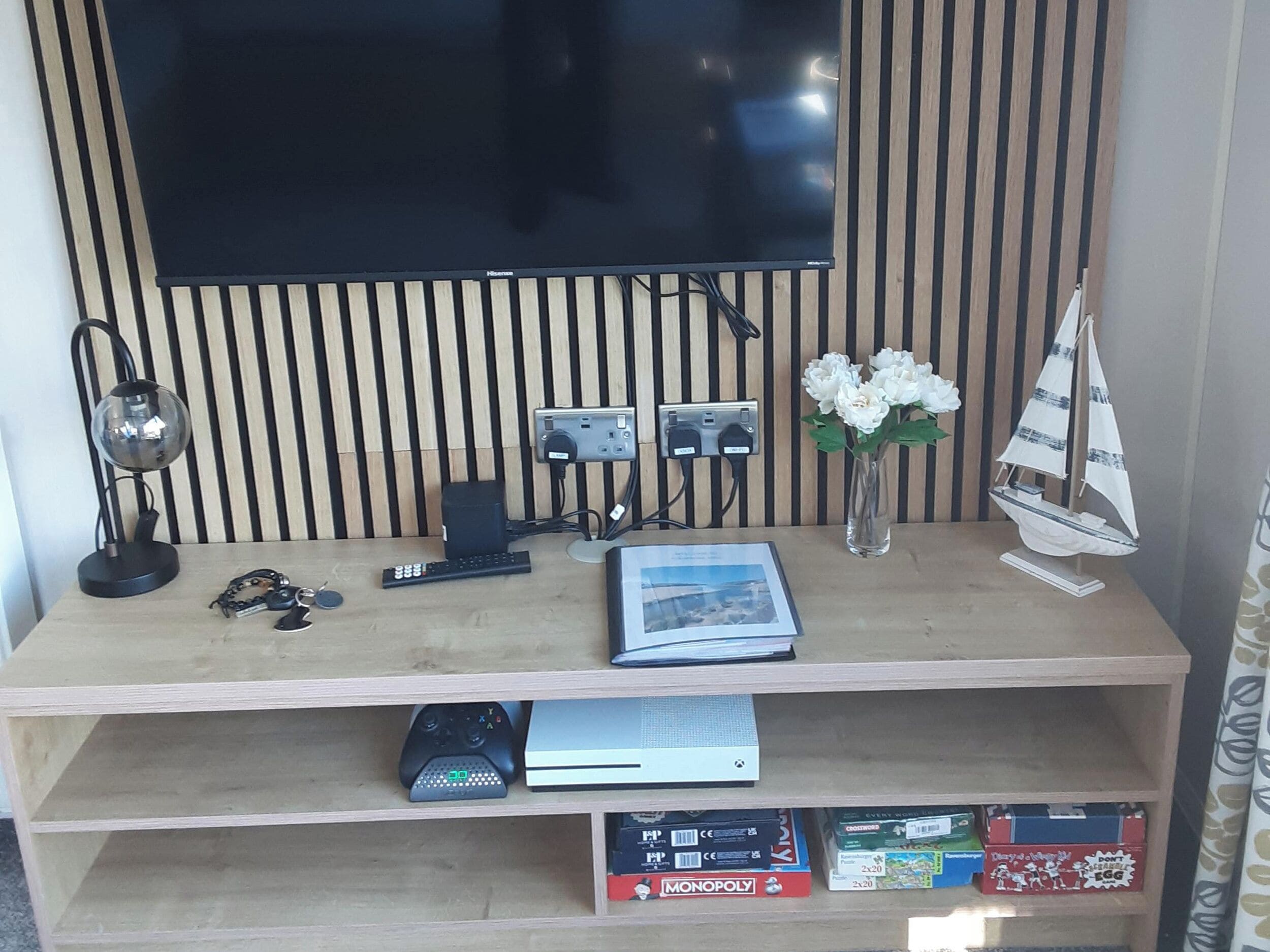 Un escritorio de madera con un televisor de pantalla plana, una lámpara, un control remoto, un florero con flores y un velero modelo.