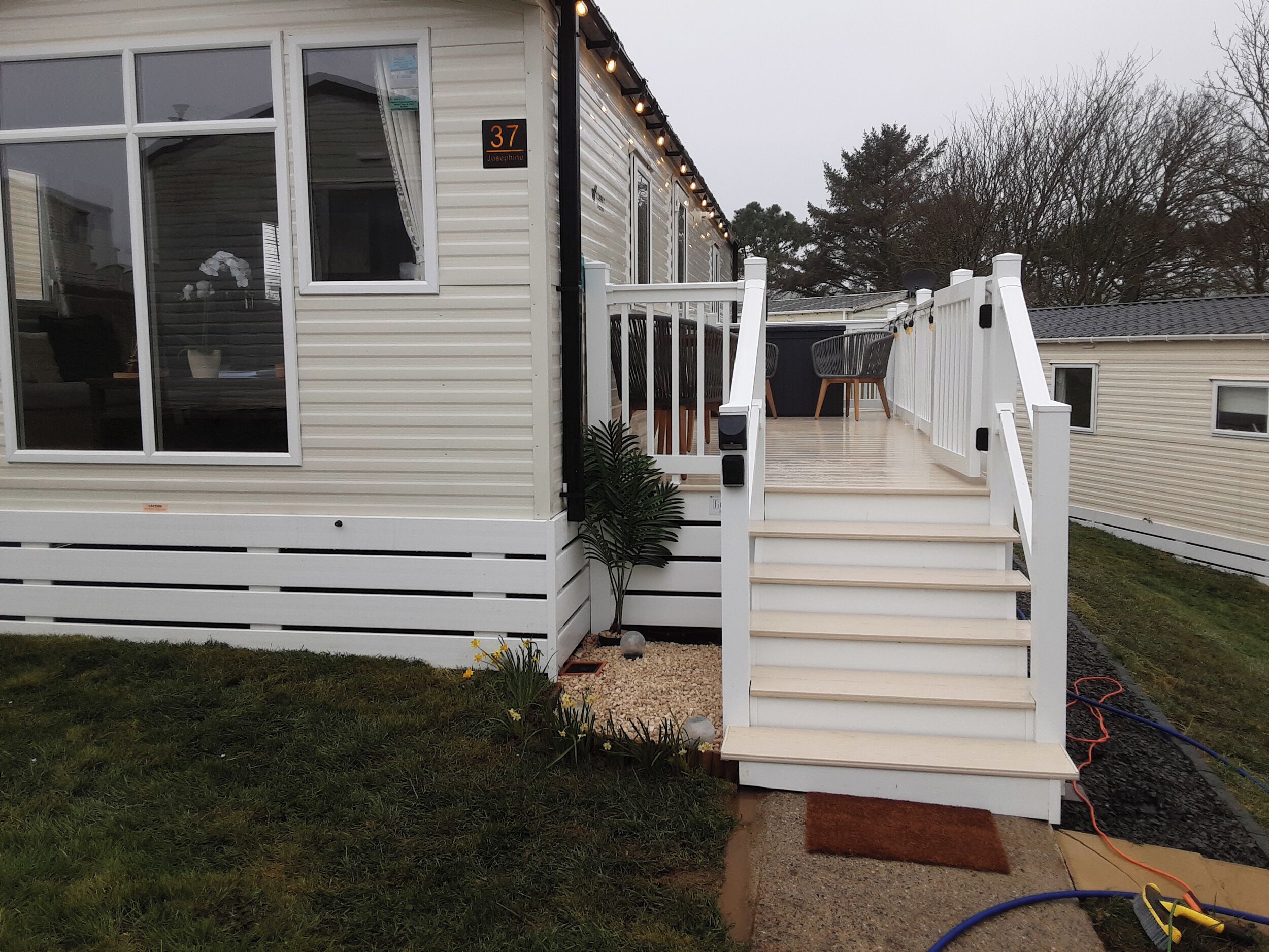 A mobile home with a white exterior, a number '37' sign, a small porch with steps, and a garden area with grass and a plant.