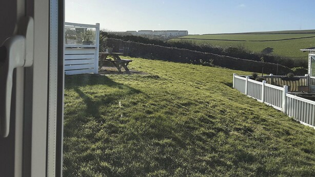 A view through a window of a grassy outdoor area with a white fence, a wooden bench, and a building in the distance. - Josephine Nb37 (Newquay)