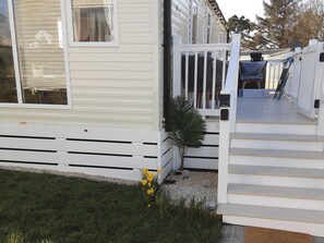 A mobile home with a white exterior, a front porch with a white railing, and a small garden area with grass and a plant. - Josephine Nb37 (Newquay)