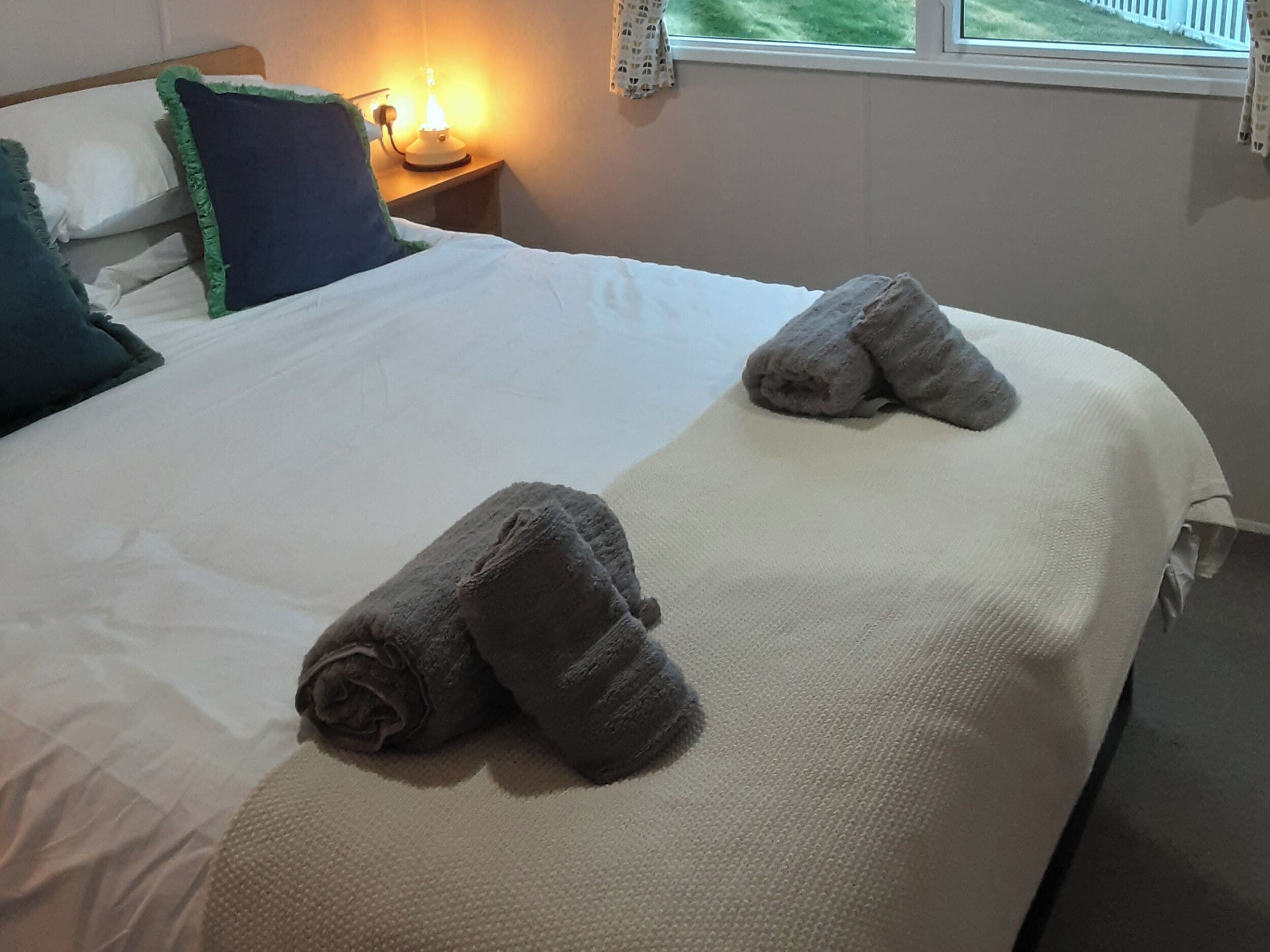 A neatly made bed with two rolled towels, a bedside lamp, and a window with a view.