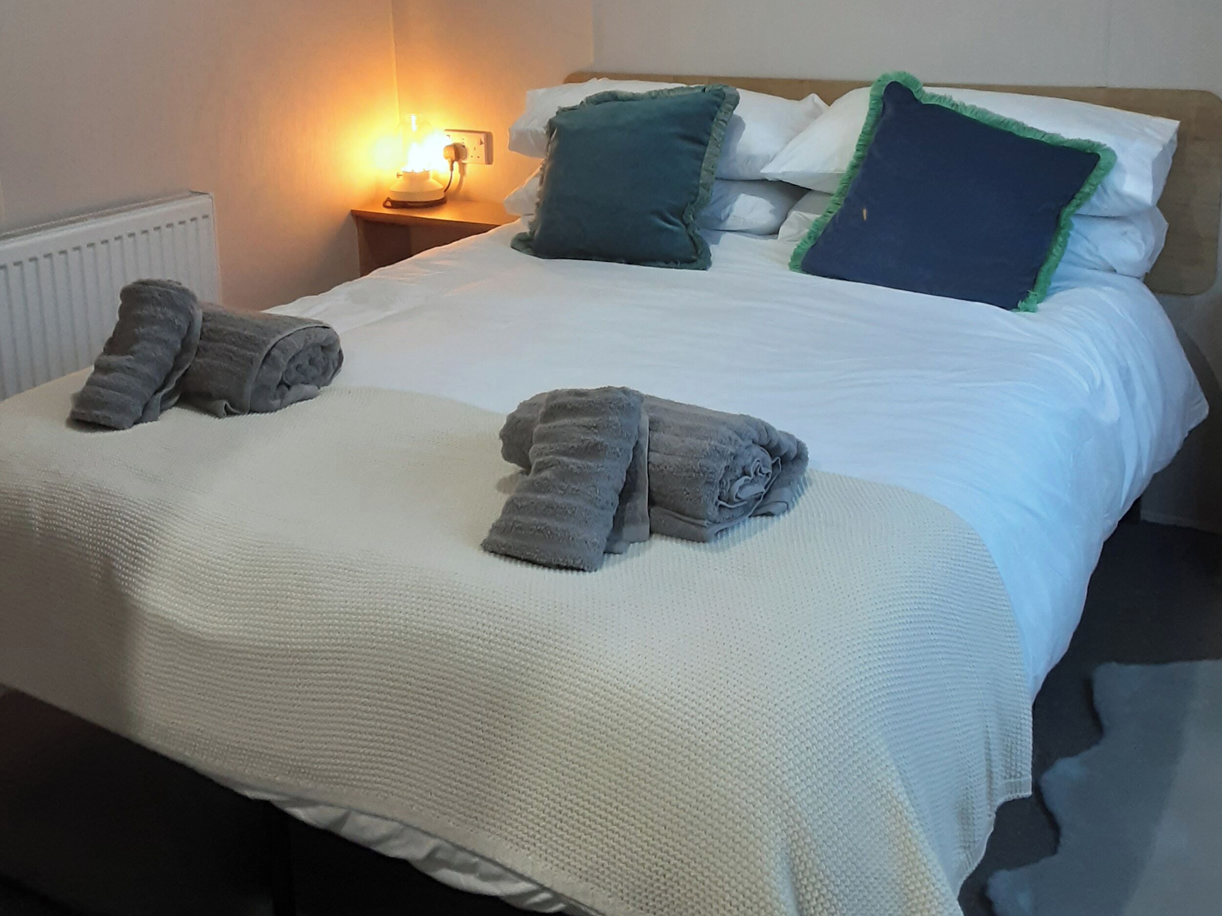 A neatly made bed with white linens, a grey blanket, and two rolled-up towels.