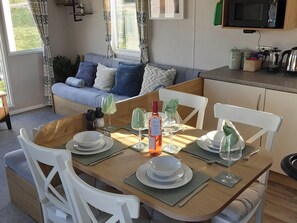 A dining area with a wooden table set for four, white chairs, and a sofa in the background. - Josephine Nb37 (Newquay)