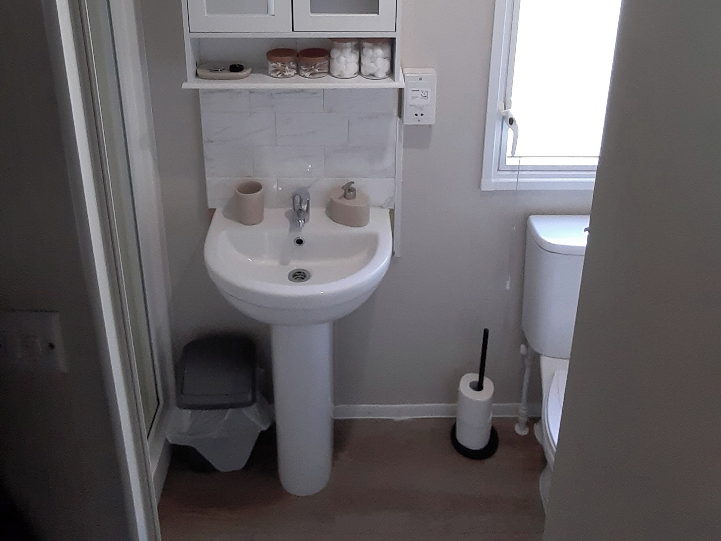 A bathroom with a pedestal sink, toilet, and a trash bin. There are storage shelves above the sink and a window to the right.