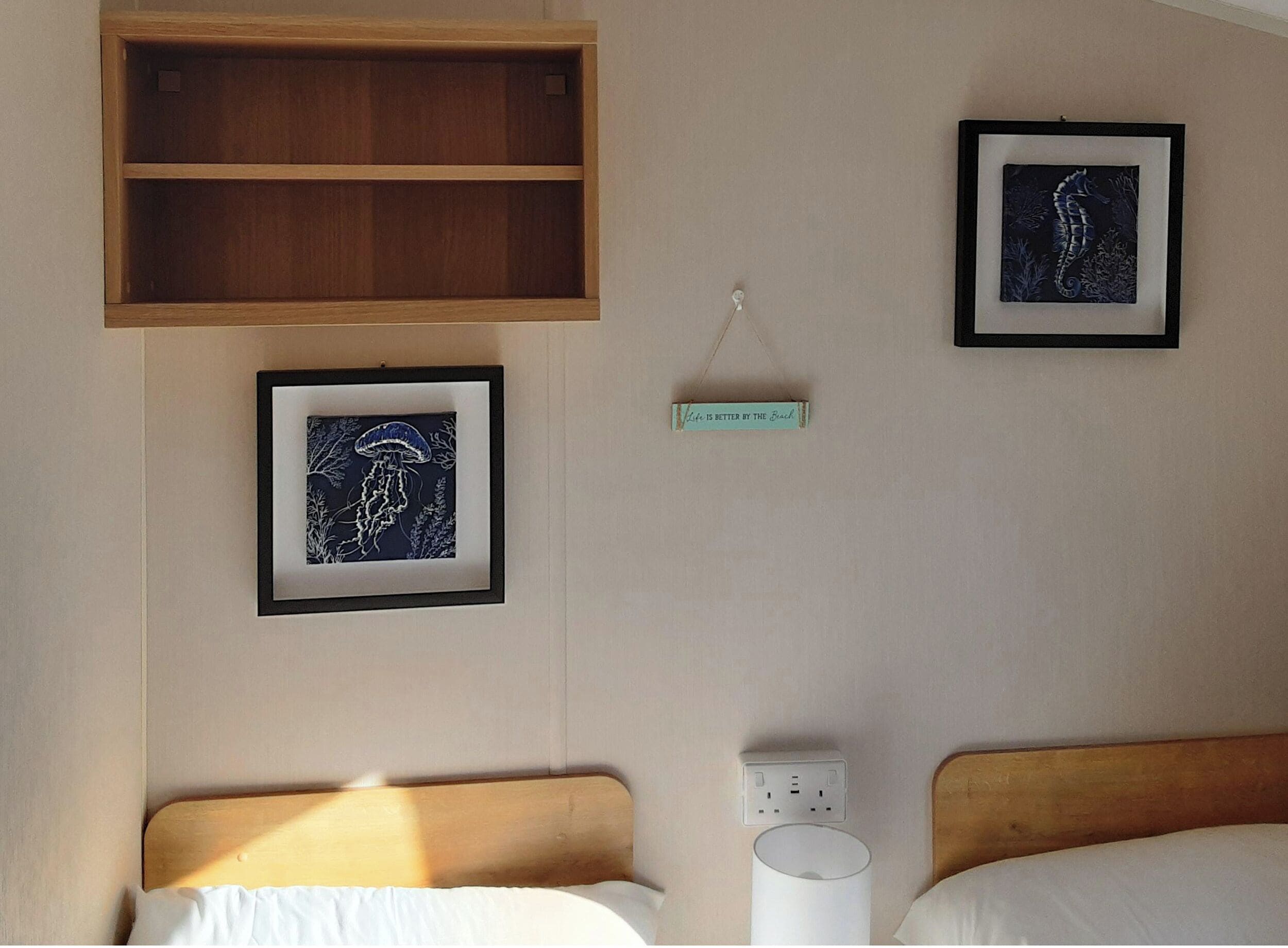 A hotel room with two beds, a wooden shelf, framed artwork, a wall socket, and a sign with text.