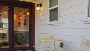 A wooden deck with a folding table and chairs, a glass door entrance, and a wall-mounted light fixture. - Family-Friendly 3 Bedroom/2 Bath Home - 25 Min to Dallas w/ Free EV Charging (Forney)
