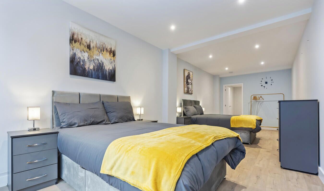 A modern bedroom with a large bed, a smaller bed, bedside tables, a wardrobe, and a clock on the wall.