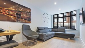 Uma sala de estar moderna com um sofá seccional, uma mesa de jantar com cadeiras, um relógio e uma janela grande com vista para edifícios.