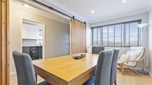 A modern living room with a wooden dining table, grey upholstered chairs, a sofa with white cushions, a large window with blinds, and a kitchen area with white cabinets.