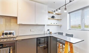 Une cuisine moderne dotée d’un lave-vaisselle encastré, d’un plan de travail avec une machine à café et d’une fenêtre avec des stores.