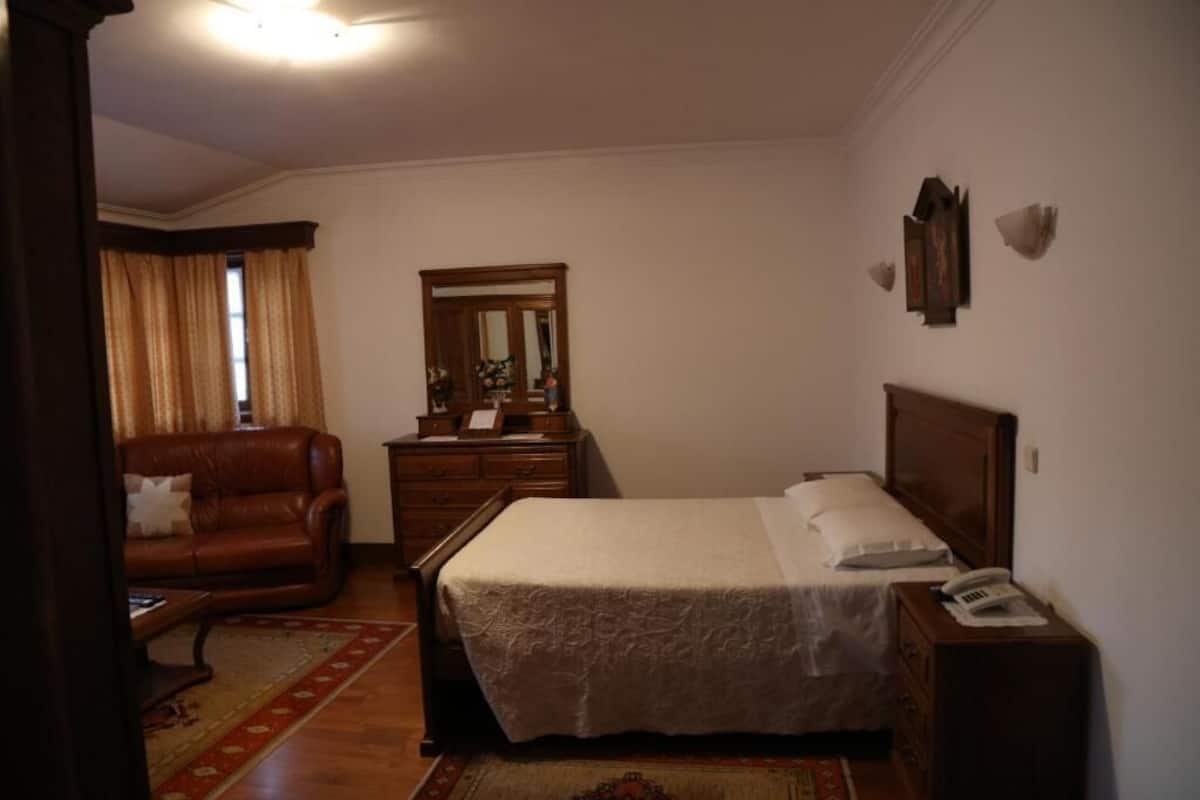 A bedroom with a bed, a dresser, a nightstand, a mirror, a window with curtains, and a leather armchair.