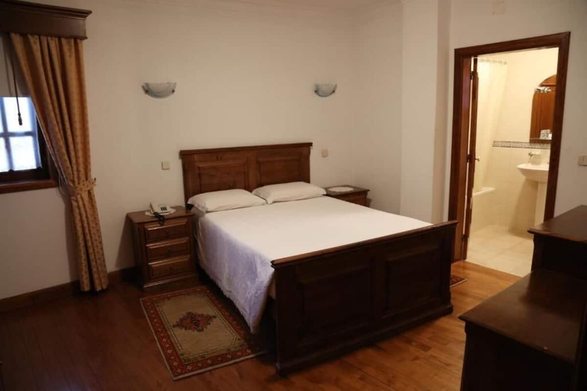 A bedroom with a wooden bed, bedside tables, a nightstand, a window with curtains, and a door leading to a bathroom.