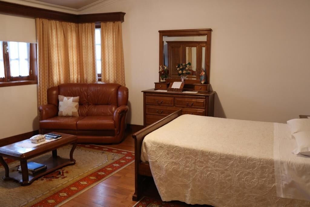 A room with a leather sofa, a wooden dresser, a bed with a white bedspread, and a small table with a remote control.