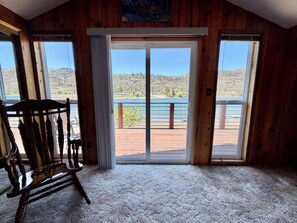 Une pièce au décor intérieur en bois, avec un fauteuil à bascule et une grande porte-fenêtre coulissante donnant sur une terrasse d’où l’on peut admirer un lac et des montagnes.