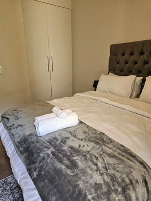 A neatly made bed with a grey and white patterned bedspread, a rolled white towel on the bed, and a dark tufted headboard. - Exquisite one-bedroom Apartment In Fourways (Sandton)
