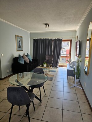 A living room with a glass-top dining table, a sofa, and a chair. - Exquisite one-bedroom Apartment In Fourways (Sandton)