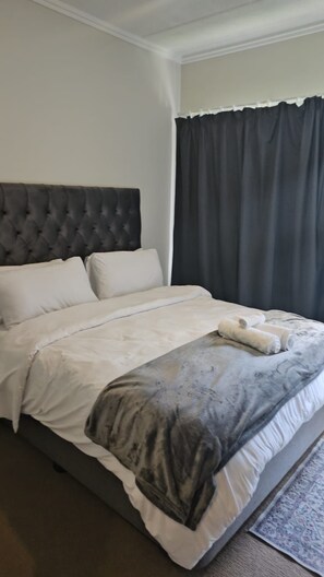 A neatly made bed with white and grey bedding, a grey headboard, and a dark curtain partially drawn. - Exquisite one-bedroom Apartment In Fourways (Sandton)