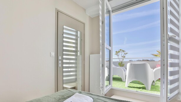 A bedroom with a bed, a window with blinds, and a view of outdoor chairs. - Appartement avec terrasse ensoleillée sur le port Olona (Les Sables-d'Olonne)